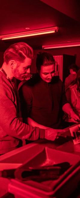 Lecturer guiding students through the film developing process under red darkroom lighting. Lecturer guiding students through the film developing process under red darkroom lighting.